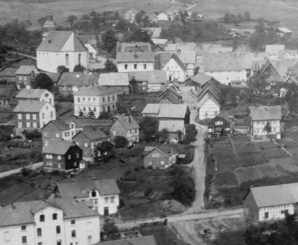 Auf diesem historischen Foto ist in der Bildmitte noch die den Achenbach’schen Scheunen gegenüberliegende Scheunengruppe erkennbar. Sie brannte in der Nacht des 12. März 1897 vollständig ab. Dieses Großfeuer gab den letzten Anstoss zur Gründung der Freiwilligen Feuerwehr Freudenberg am 4. Mai 1897. Die Krottorferstraße bildete damals fast eine Einheit mit der Bergstraße, im Volksmund „Holle“ genannt. Ebenso lässt diese Aufnahme deutlich die Rückseiten der Scheunenreihe unterhalb von Pfarrhaus und Kirche erkennen. 